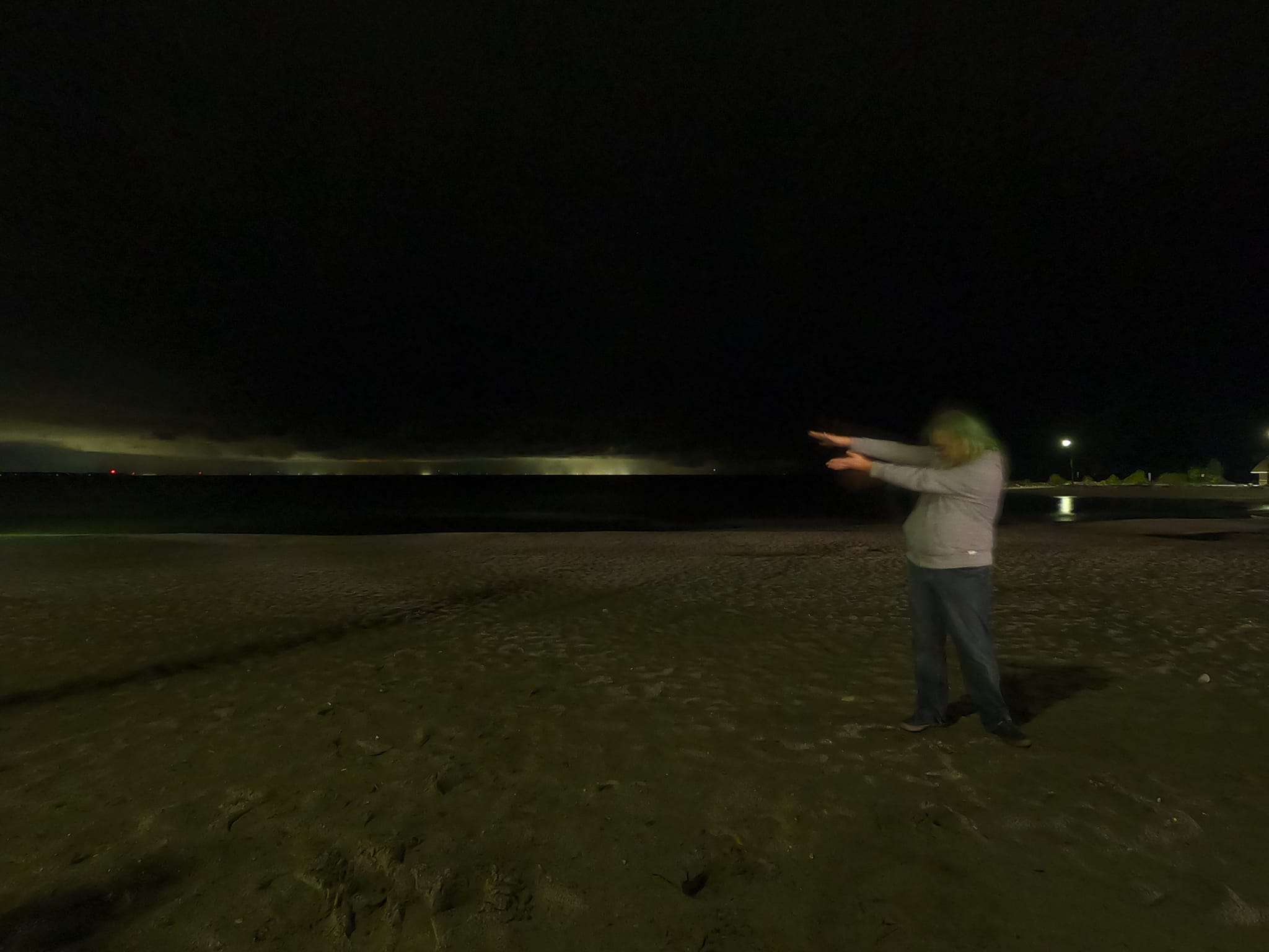 A man with green hair poses on a beach at night with the lights across the bay